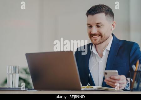 Businessman using laptop and smartphone with earphones while working online Stock Photo