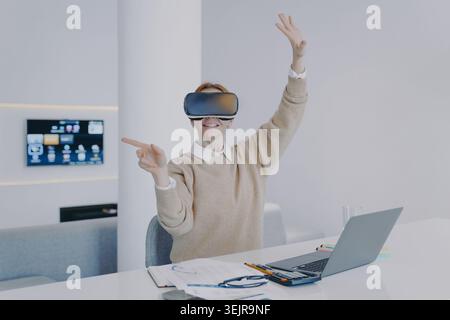 Business woman using virtual reality headset at desk with laptop in modern office Stock Photo