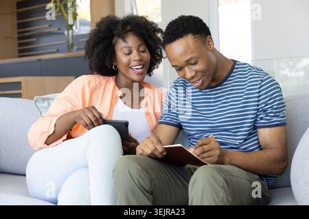 African American couple sitting on gray sofa in living room, holding tablet and writing notebook. Duo, collaboration, lounge, mo Stock Photo