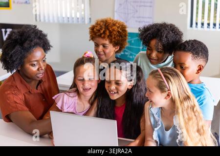 Diverse classroom teacher guiding school-age children clustering around laptop on white desk. Collaborative, learning, instructi Stock Photo