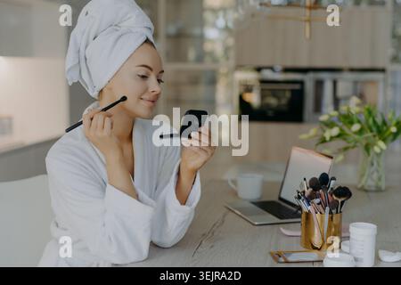 Woman applying makeup at home in bathrobe with towel on head Stock Photo