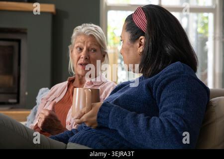 Senior female friends chatting while holding ceramic mugs on living room sofa near fireplace. Cozy, home, duo, conversation, war Stock Photo