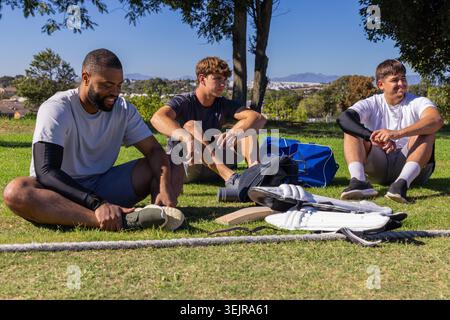Three adult men sitting on grassy field, resting and checking cricket bat, pads, bag. Outdoor, park, teammates, camaraderie, sportswear, equipment, su Stock Photo