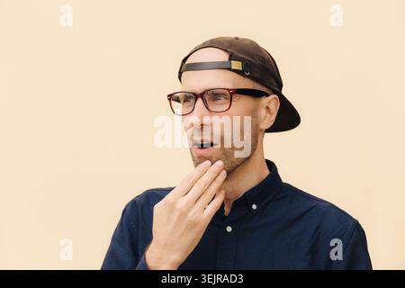 Surprised man in glasses and cap touching chin with shocked expression Stock Photo