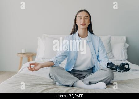Young woman with prosthetic arm meditating on bed in bedroom at home Stock Photo