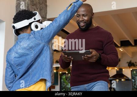 African American males in kitchen, adult in sweater guiding denim-jacket teen using VR and tablet. Family, home, cozy, warm, hea Stock Photo