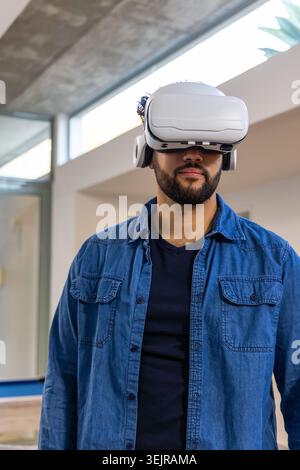 Man wearing white VR headset and open denim shirt, standing in modern office with skylight. Wearable, goggles, technology, immer Stock Photo
