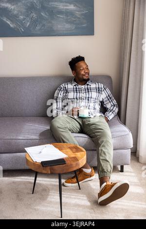 African American man in plaid shirt and joggers sitting on gray sofa at home holding mug. Serene, cozy, minimalist, natural, lig Stock Photo