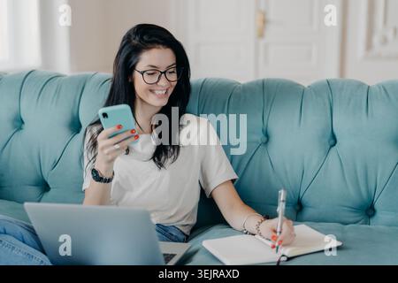 Woman checking smartphone and writing notes on sofa while working from home Stock Photo