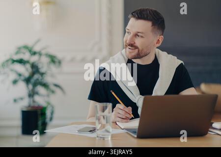 Young man writing notes while using laptop at home office desk Stock Photo