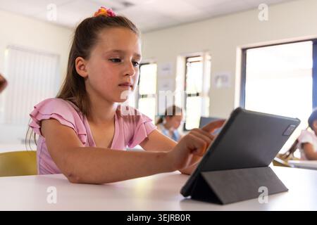 School-age girl in pink ruffle top sitting at school desk tapping tablet in dark folio case. Classroom, student, digital, learning, daylight, focus, i Stock Photo