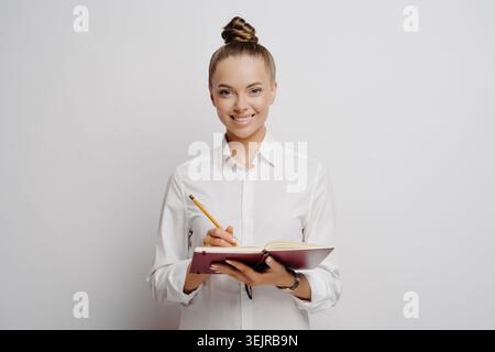 Smiling Young Woman Taking Notes in Notebook with Pencil on Clean White Background Stock Photo