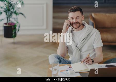 Casual Man Talking on Smartphone and Writing Notes at Home Desk with Laptop and Papers Stock Photo