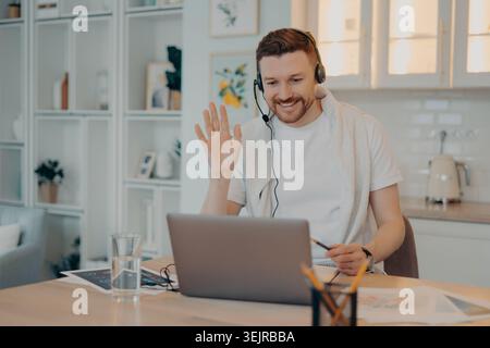 Young Man Wearing Headset Using Laptop at Home Office Waving During Online Video Call Stock Photo