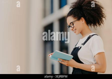 Young Woman Holding Notebook Outside Modern Building Smiling While Reading Notes in Daylight Stock Photo