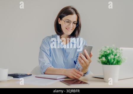 Woman Using Smartphone at Desk While Writing Notes in Bright Modern Home Office Stock Photo