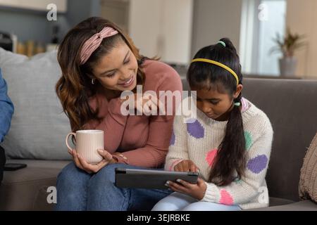 Diverse mother and child sitting on gray sofa at home, mother holding mug, child holding tablet. Parent, daughter, screen, device, family, cozy, warm Stock Photo