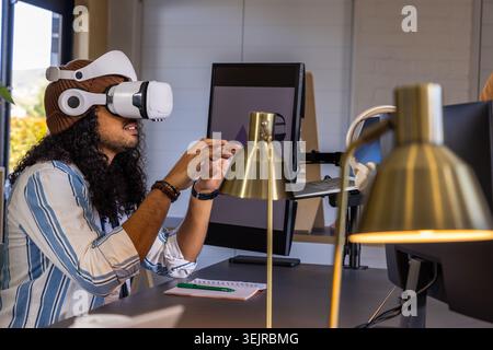 Adult male in striped shirt and beanie using white VR headset manipulating virtual objects at desk. Workspace, office, monitors, Stock Photo