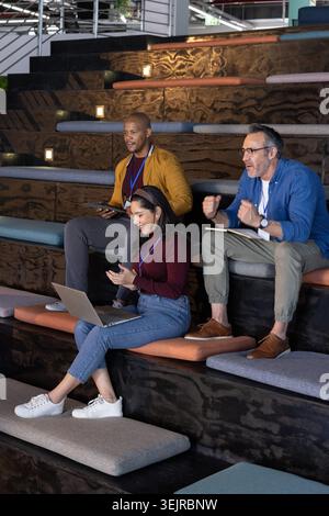 Diverse coworkers collaborating in amphitheater office wearing business clothes with laptop, tablet. Collaboration, colleagues, coworking, notebook, d Stock Photo