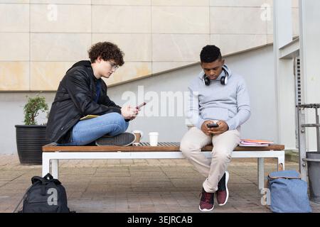 Diverse teen male students sitting on bench in campus plaza using smartphones with coffee cups. Courtyard, urban, outdoor, duo, Stock Photo