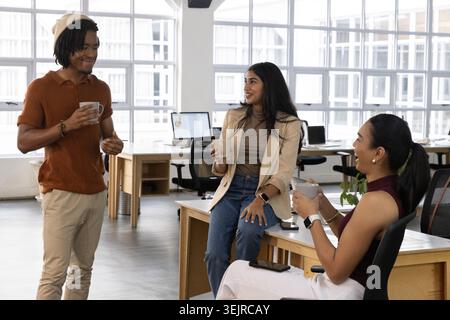 Three colleagues sharing coffee, man raising mug starting casual chat in flat animated office. Daylight, openplan, collaboration, teamwork, windows, m Stock Photo