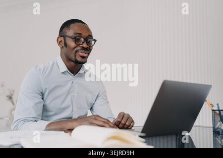 Focused Black Professional Man Working on Laptop at Desk in Bright Home Office with Notebook and Papers Stock Photo