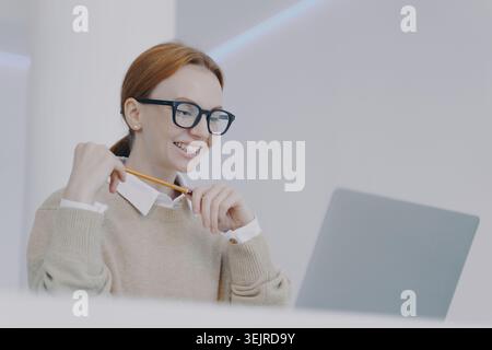 Focused Red Haired Woman Using Laptop and Holding Pencil in Modern Office During Online Work or Study Stock Photo