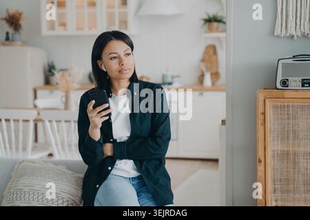 Pensive Young Woman Using Smartphone at Home Listening to Earbuds and Reading Message Stock Photo