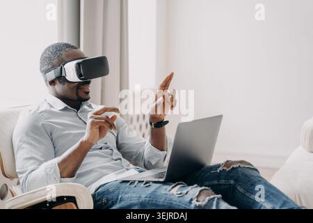 Curious Black Man Wearing Virtual Reality Headset While Using Laptop at Home in Bright Living Room Stock Photo