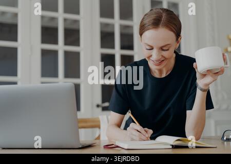 Focused Woman Taking Notes at Home Desk with Laptop and Coffee During Remote Work Stock Photo