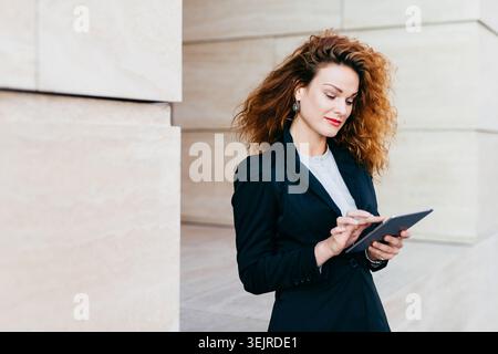 Focused Professional Woman Using Digital Tablet in Modern Office Corridor Checking Work Information on Touchscreen Stock Photo