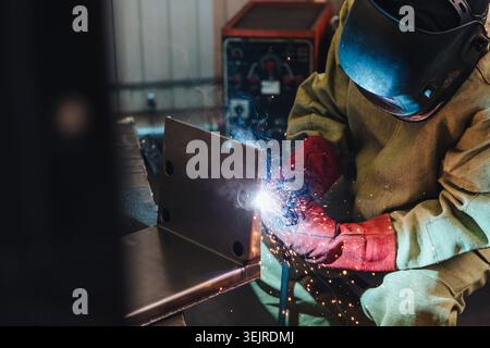 Focused Welder Joining Metal Frame in Industrial Workshop with Bright Arc Sparks, Protective Helmet, Red Gloves, and Heavy Equip Stock Photo