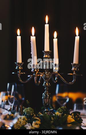 Elegant Silver Candelabra with Burning Candles on Formal Dinner Table in Dark Restaurant Interior with Flowers and Wine Glasses Stock Photo