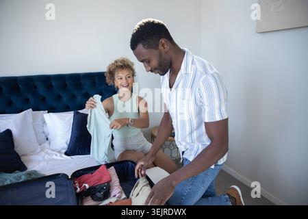 Diverse couple sitting on bed edge holding striped shirt and packing suitcase in bedroom. Modern, cozy, vibrant, lifestyle, relaxation, intimacy, casu Stock Photo