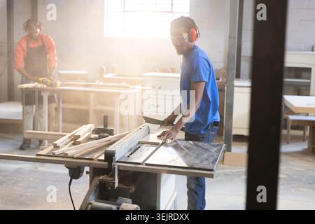 African American male coworkers guiding plank through table saw using drill in carpentry workshop. Craftsmanship, woodworking, i Stock Photo
