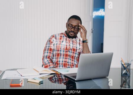 Thoughtful Man Working on Laptop at Office Desk Reviewing Documents and Notes with Serious Expression in Modern Workspace Stock Photo