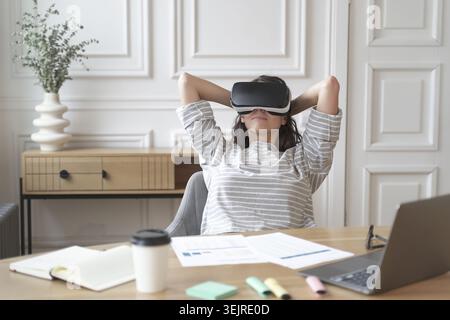 Relaxed woman wearing virtual reality headset leaning back at home office desk during break from laptop work and paperwork Stock Photo