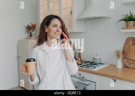 Smiling pregnant woman talking on smartphone and holding takeaway coffee in bright home kitchen during calm morning routine Stock Photo