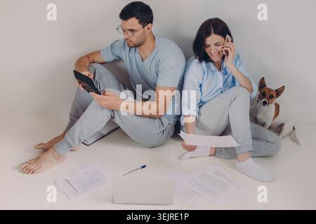 Relaxed Young Couple Managing Household Bills on Floor with Laptop Tablet Phone and Small Dog in Minimal Home Interior Stock Photo