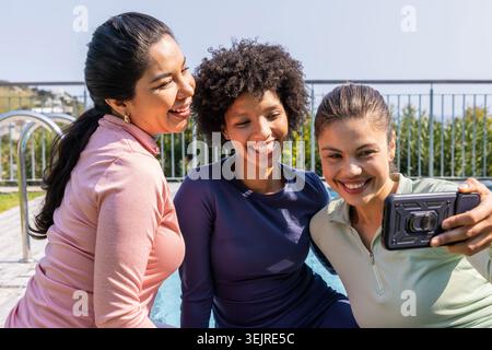 Diverse female friends posing for selfie poolside in long-sleeve athletic tops holding smartphone. Trio, rooftop, deck, outdoors, sunny, water, active Stock Photo