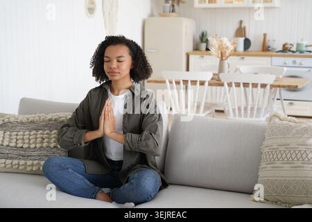 Calm Young Black Woman Meditating on Sofa at Home with Hands in Prayer Pose Practicing Mindfulness and Breathing Exercise Stock Photo