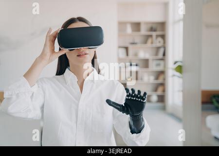 Curious Young Woman Wearing Virtual Reality Headset at Home While Using Futuristic Prosthetic Hand in Bright Modern Room Stock Photo