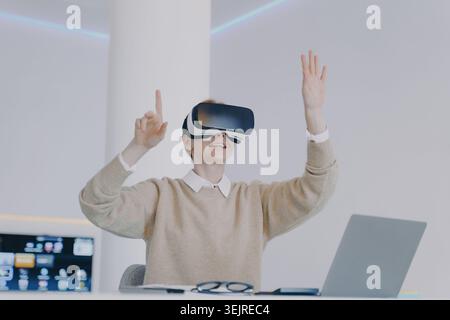 Excited Woman Wearing Virtual Reality Headset at Desk Using Laptop and Raising Hands in Bright Modern Home Office Stock Photo