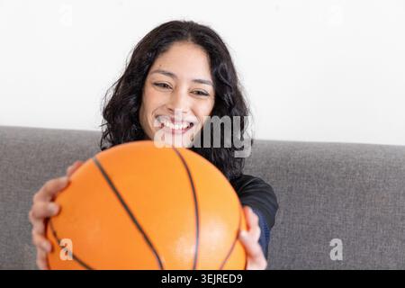Female sitting on grey sofa holding orange basketball toward camera while smiling, wearing dark top. Ball, woman, home, curly, c Stock Photo