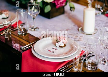 Elegant Wedding Table Setting with White Plates Red Napkin Candle Glassware and Small Guest Favor on Rustic Reception Table Stock Photo