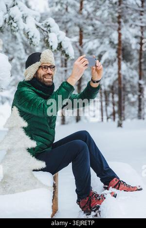 Bearded man uses smartphone while sitting in snowy forest during winter hike Stock Photo