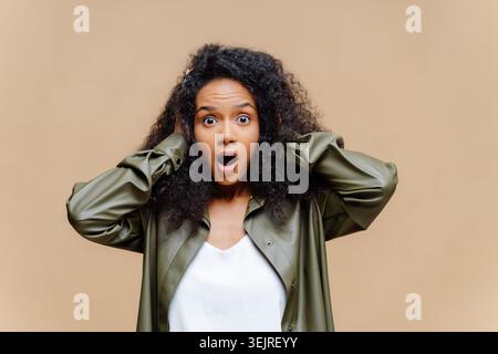 Surprised young woman holds her head while posing against beige studio wall Stock Photo