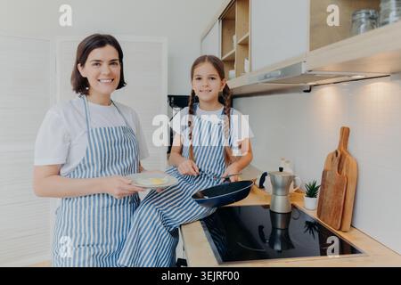 Mother and daughter wearing striped aprons cook pancakes together in home kitchen Stock Photo