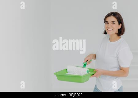 Young woman holds paint roller and tray while renovating white room at home Stock Photo