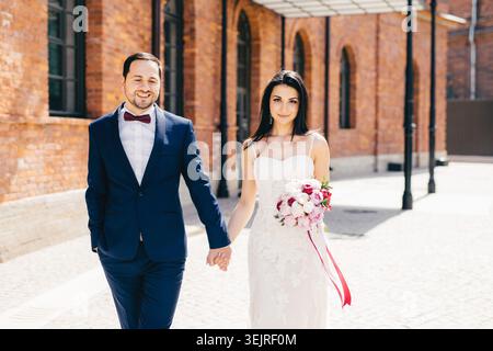Bride and groom walking hand in hand with bouquet near brick wedding venue Stock Photo